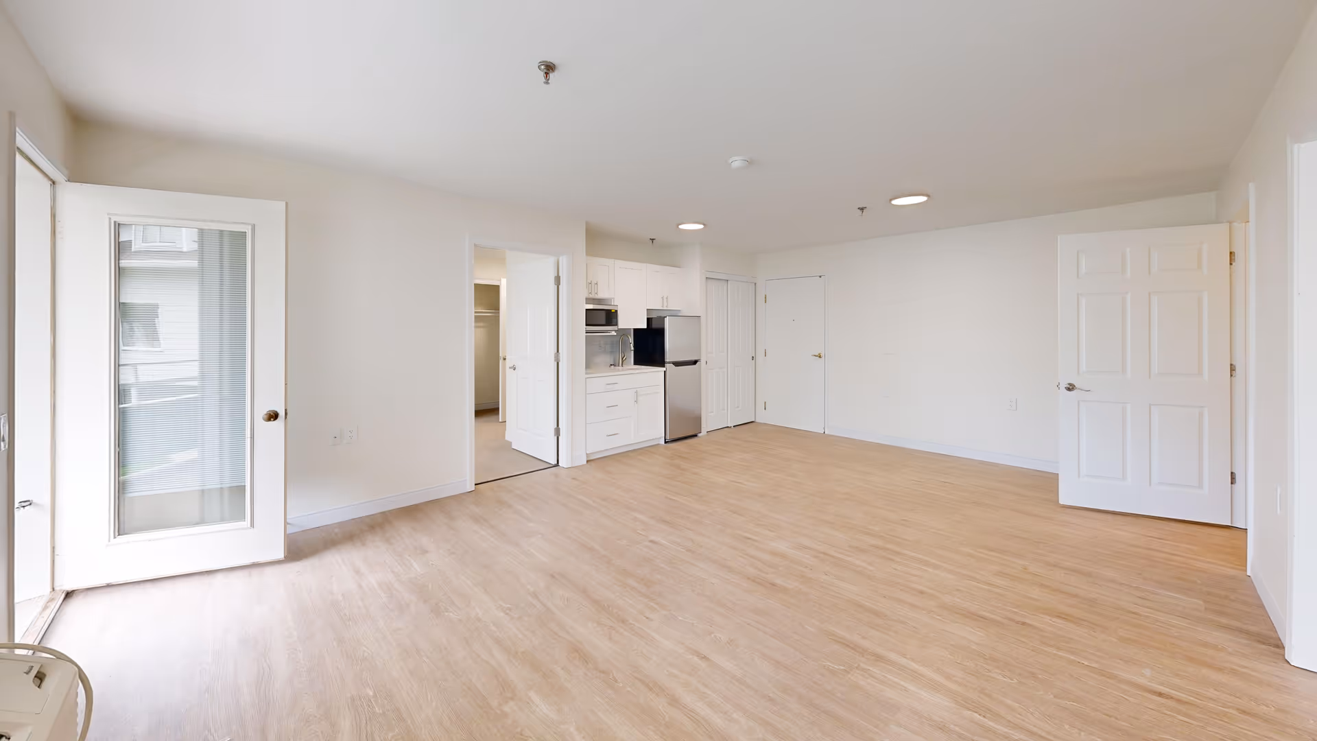 A bright, empty apartment room with light wood flooring, white walls, and a small kitchen area with white cabinets, a microwave, and a refrigerator. There are two open doors leading to other rooms and a glass door opening to a balcony or outdoor area.