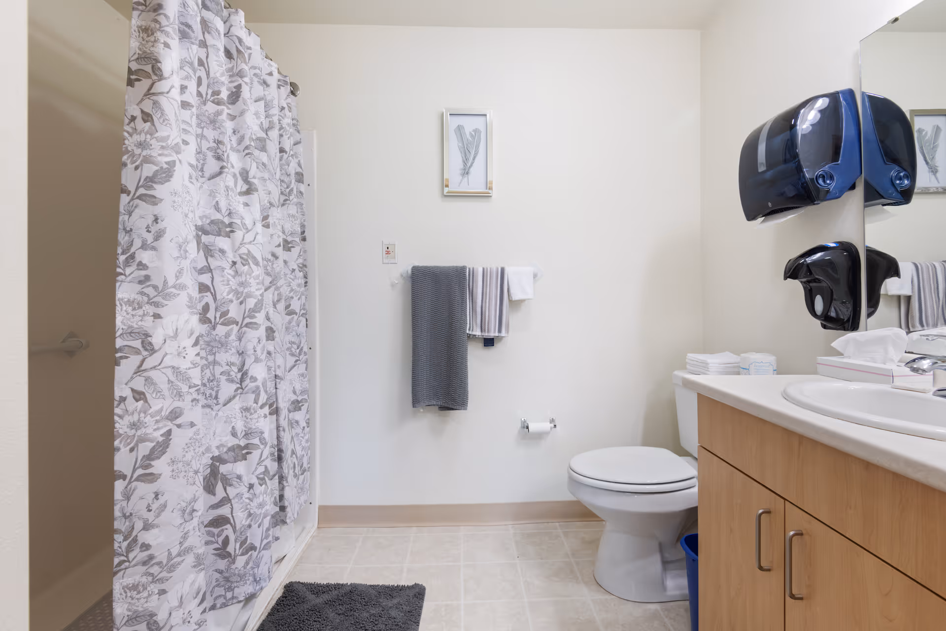 A clean senior living facility bathroom featuring a shower with a floral patterned curtain, a toilet, a sink with a countertop and wooden cabinets, a wall-mounted soap dispenser, a hand dryer, a towel rack with two towels, a small framed picture on the wall, and a dark gray bath mat on the floor.