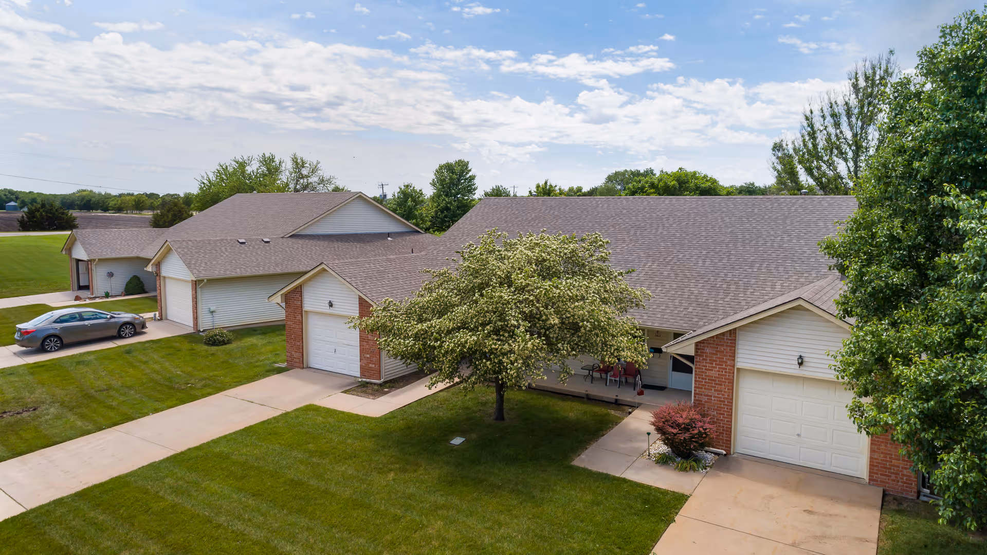 Exterior view of a senior living facility showing multiple single-story buildings with garages, well-maintained green lawns, a tree in the center, and a partly cloudy sky.