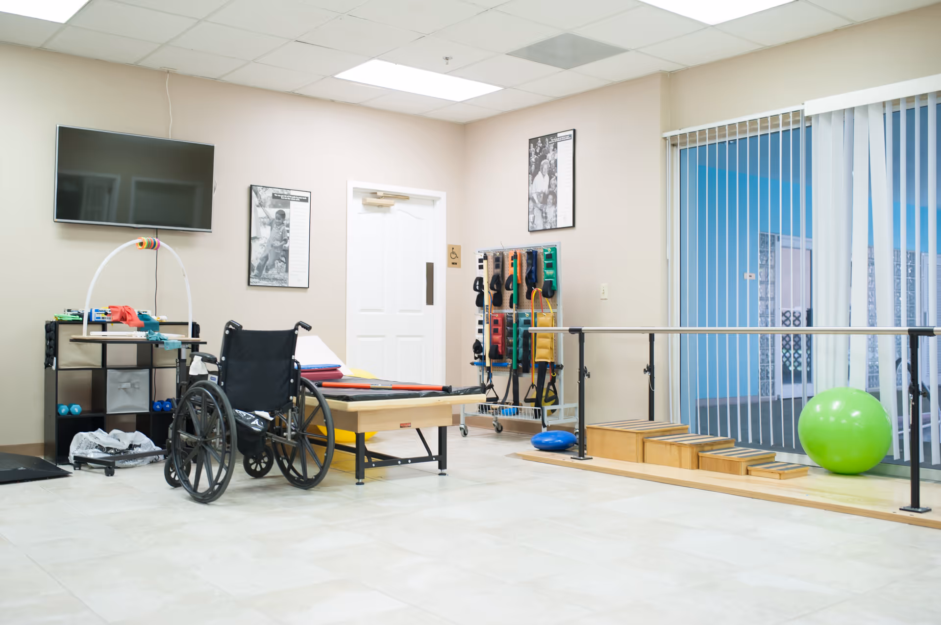 A rehabilitation or physical therapy room with a wheelchair, therapy bed, exercise equipment including resistance bands on a rack, a green exercise ball, and parallel bars with wooden steps. The room has beige walls, a mounted flat-screen TV, and vertical blinds covering a large window.