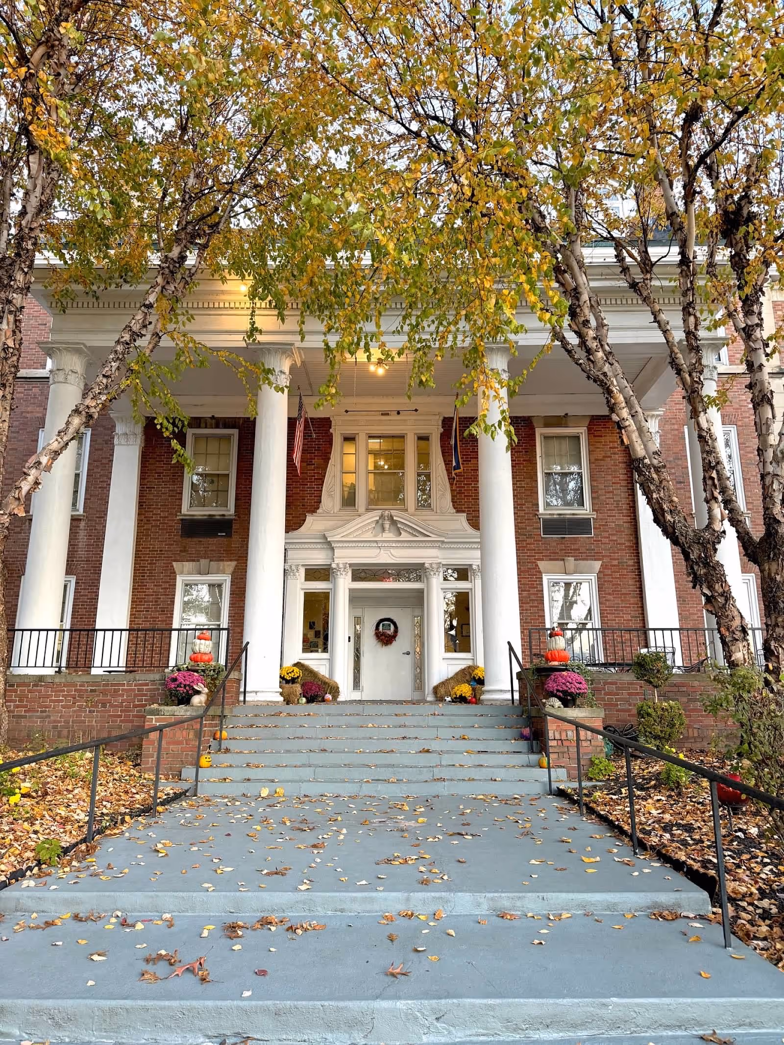Front entrance of a brick building with white columns and a wide staircase leading up to the door. The entrance is decorated with autumn-themed flowers and pumpkins. Trees with yellowing leaves frame the view.