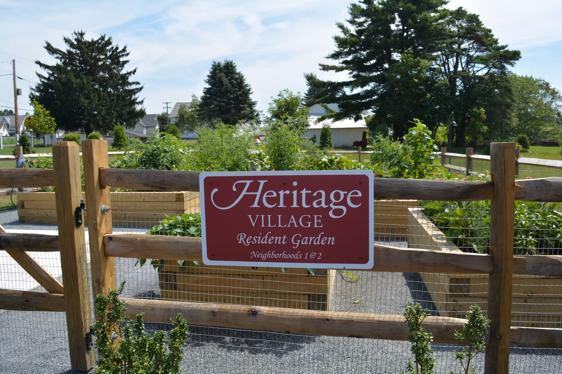 A wooden fenced garden area with raised garden beds filled with green plants. A red sign on the fence reads 'Heritage Village Resident Garden Neighborhoods 1 & 2'. Trees and houses are visible in the background under a partly cloudy sky.