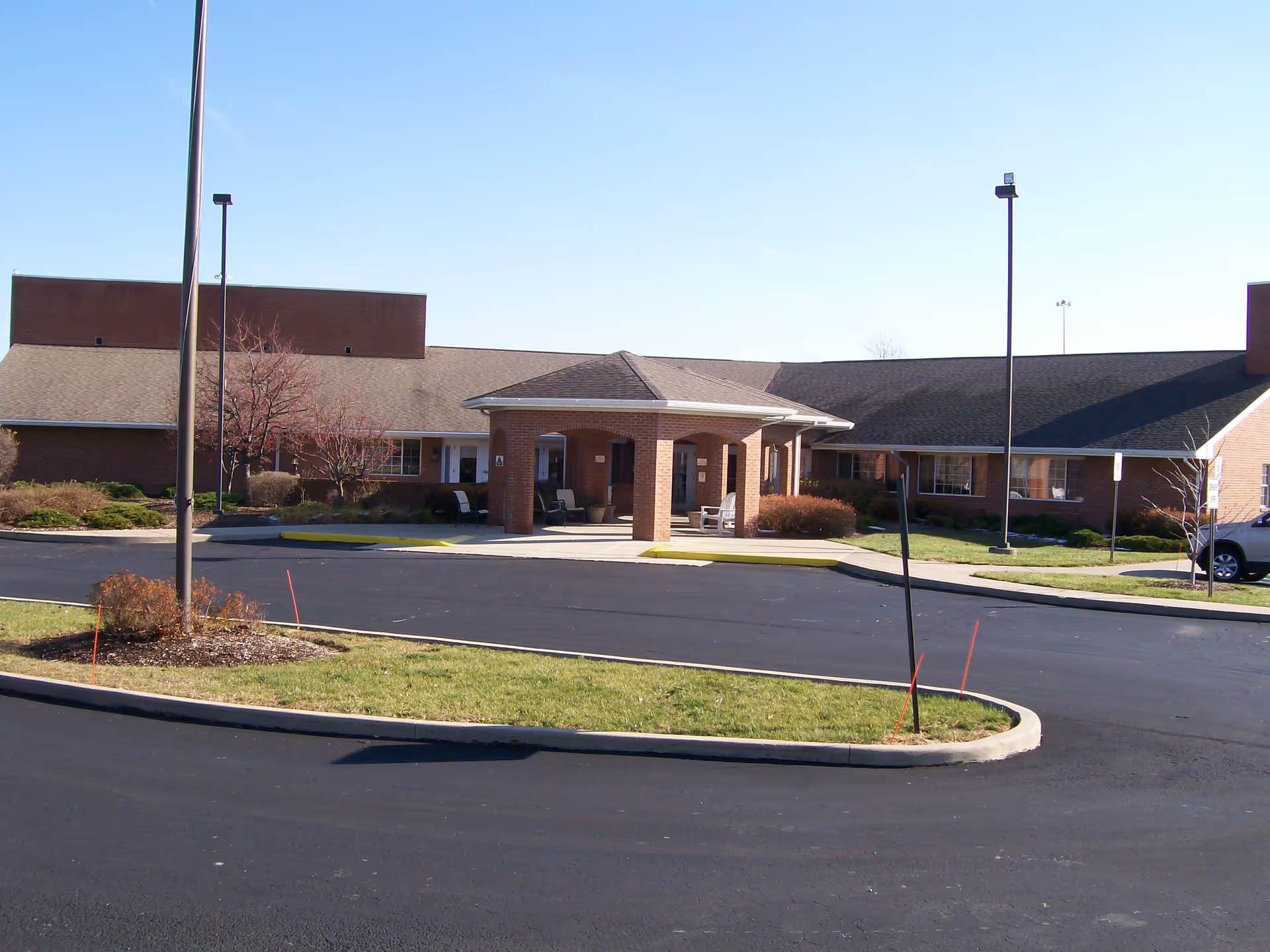 Exterior view of Manor At Perrysburg senior living facility showing a single-story brick building with a covered entrance, surrounded by a paved driveway and landscaped grass areas under a clear blue sky.