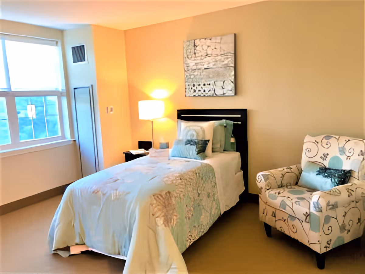 A cozy bedroom in a senior living facility featuring a single bed with light blue and beige bedding, a decorative pillow, a bedside table with a lamp, a patterned armchair with a matching pillow, and a window with blinds letting in natural light.