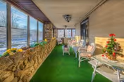 A sunroom or enclosed porch with green carpet flooring, stone half-walls, and large windows overlooking an outdoor area. The room is furnished with white wicker chairs and tables, some with cushions and small decorative items including a small Christmas tree with red ornaments.