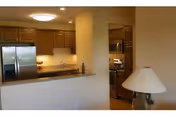 Interior view of a senior living facility kitchen and part of a living area. The kitchen features wooden cabinets, a stainless steel refrigerator, microwave, and stove. A countertop with a raised bar separates the kitchen from the living area, where a table lamp is visible.