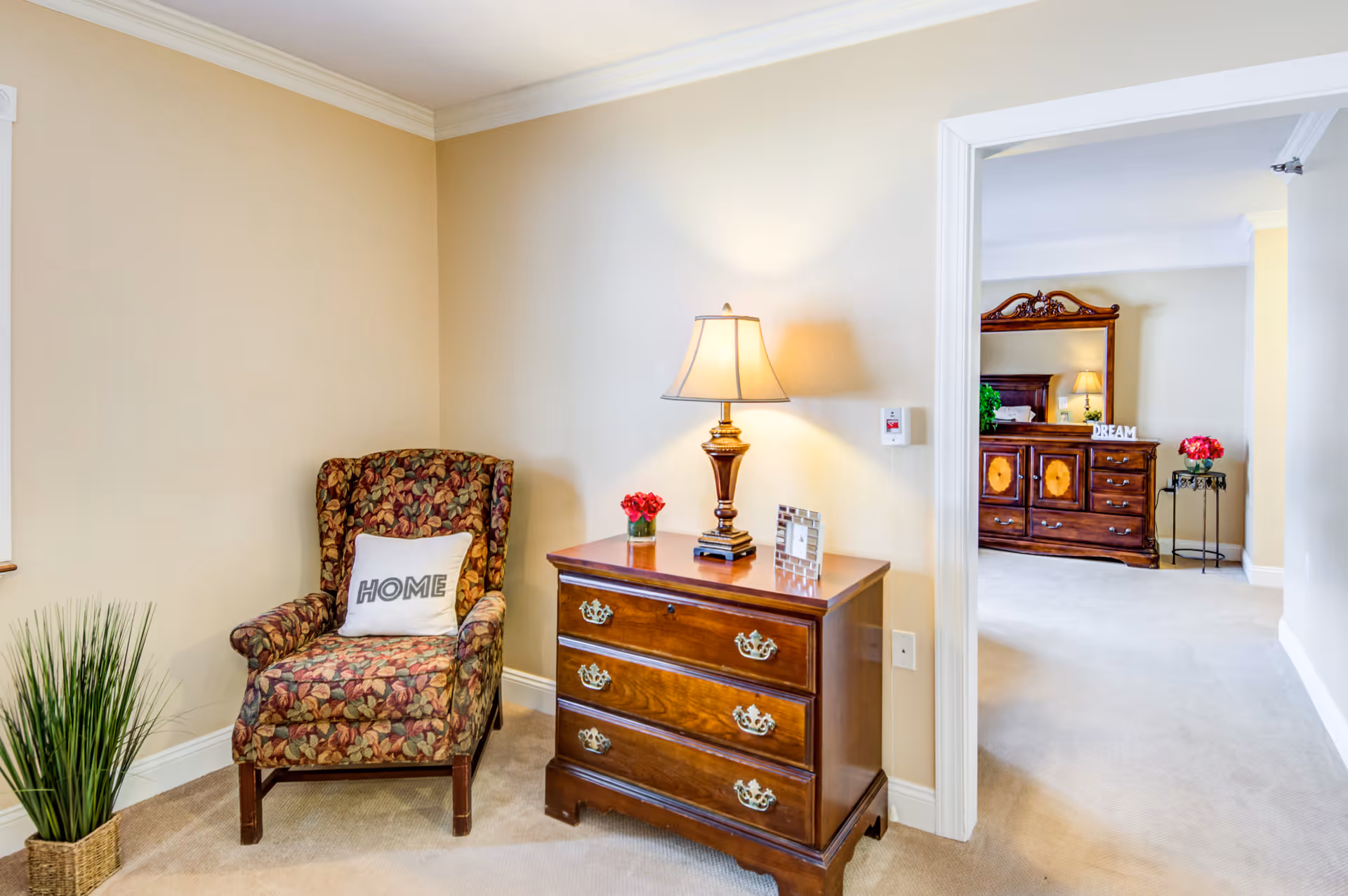 A cozy corner of a senior living facility room featuring a floral upholstered armchair with a white pillow labeled 'HOME', a wooden three-drawer dresser with a table lamp, a small vase of red flowers, and a picture frame. The room has beige walls and carpeted floors. Through an open doorway, a bedroom with a wooden dresser and mirror is visible, decorated with a lamp, a 'DREAM' sign, and another small table with red flowers.