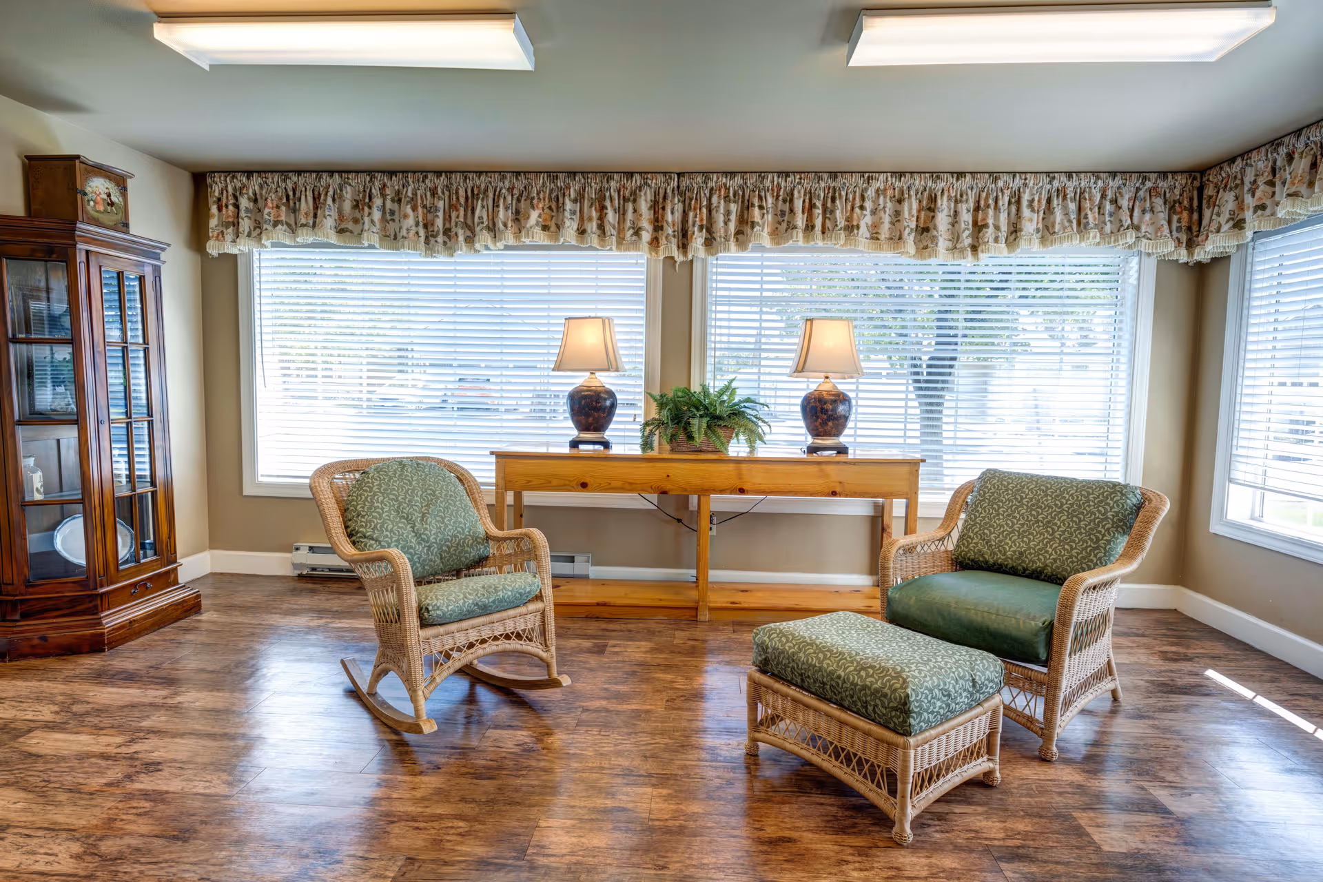 Sunlit sitting area with wicker chairs and ottoman, a wooden console table with lamps and plant, and large windows with blinds.