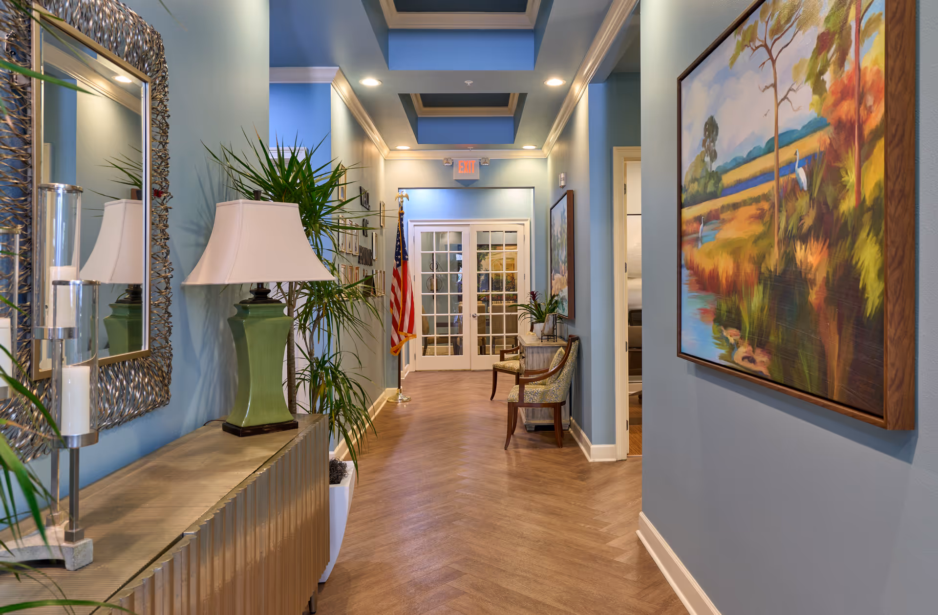 A well-lit hallway with blue walls and wooden herringbone flooring. The hallway features decorative elements including a large mirror with a textured frame, a green lamp with a white shade on a console table, potted plants, framed artwork, and two upholstered chairs near a small table. At the end of the hallway, there are glass double doors and an American flag on a stand. Recessed ceiling lights illuminate the space.