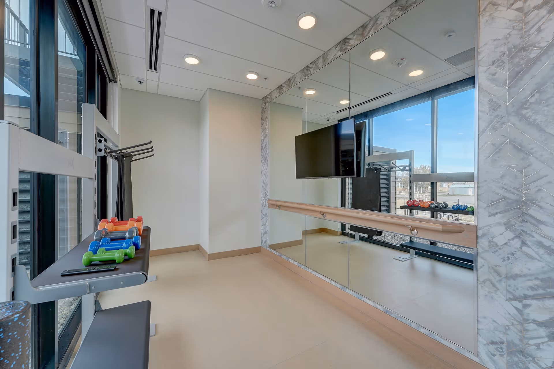 A small exercise room with a wall-mounted ballet barre and large mirrors. There is a flat-screen TV mounted on the mirrored wall. A rack with colorful dumbbells is positioned near large windows that let in natural light, and a foam roller is visible on the floor.