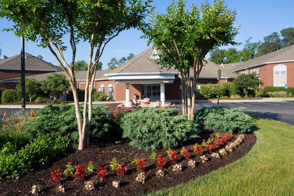 Front exterior view of Chestnut Grove Assisted Living facility with well-maintained landscaping including trees, bushes, and colorful flower beds under a clear blue sky.