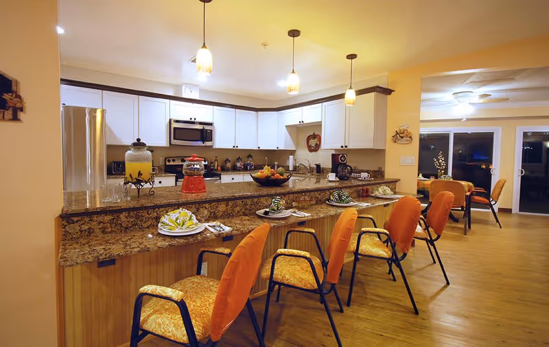 A well-lit kitchen and dining area with a granite countertop bar featuring four orange cushioned chairs. The kitchen has white cabinets, stainless steel appliances including a refrigerator and microwave, and various decorative items on the counter. In the background, there is a dining table with chairs and sliding glass doors.