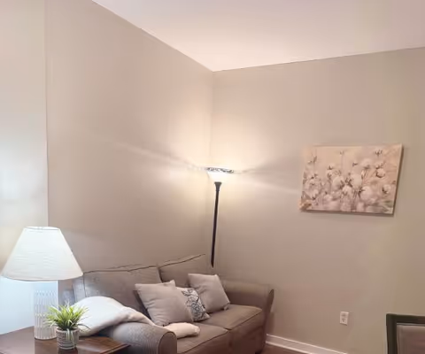 A cozy living room corner with a gray sofa adorned with several pillows and a folded blanket. There is a floor lamp in the corner emitting soft light, a table lamp on a side table with a small potted plant, and a floral painting hanging on the beige wall.
