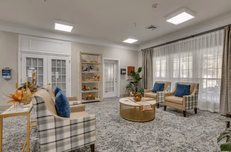 A cozy living room area with four beige and plaid armchairs, each with a blue cushion. A round coffee table with decorative items sits in the center on a patterned carpet. Large windows with sheer white curtains and beige drapes allow natural light to fill the room. A wooden shelving unit with autumn-themed decorations is against the wall near a door. The walls are light-colored, and the ceiling has recessed lighting.