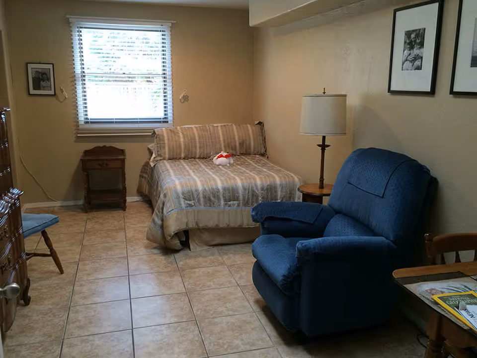 Small furnished bedroom with a bed against the back wall, a blue recliner, side table and window, on a tiled floor.