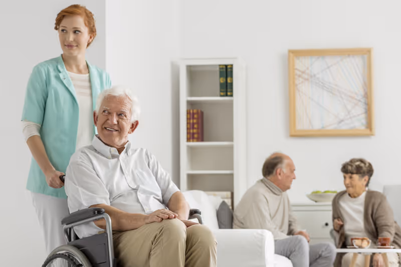 A caregiver stands behind an elderly man in a wheelchair while two other seniors sit and talk on a sofa in a bright living room.