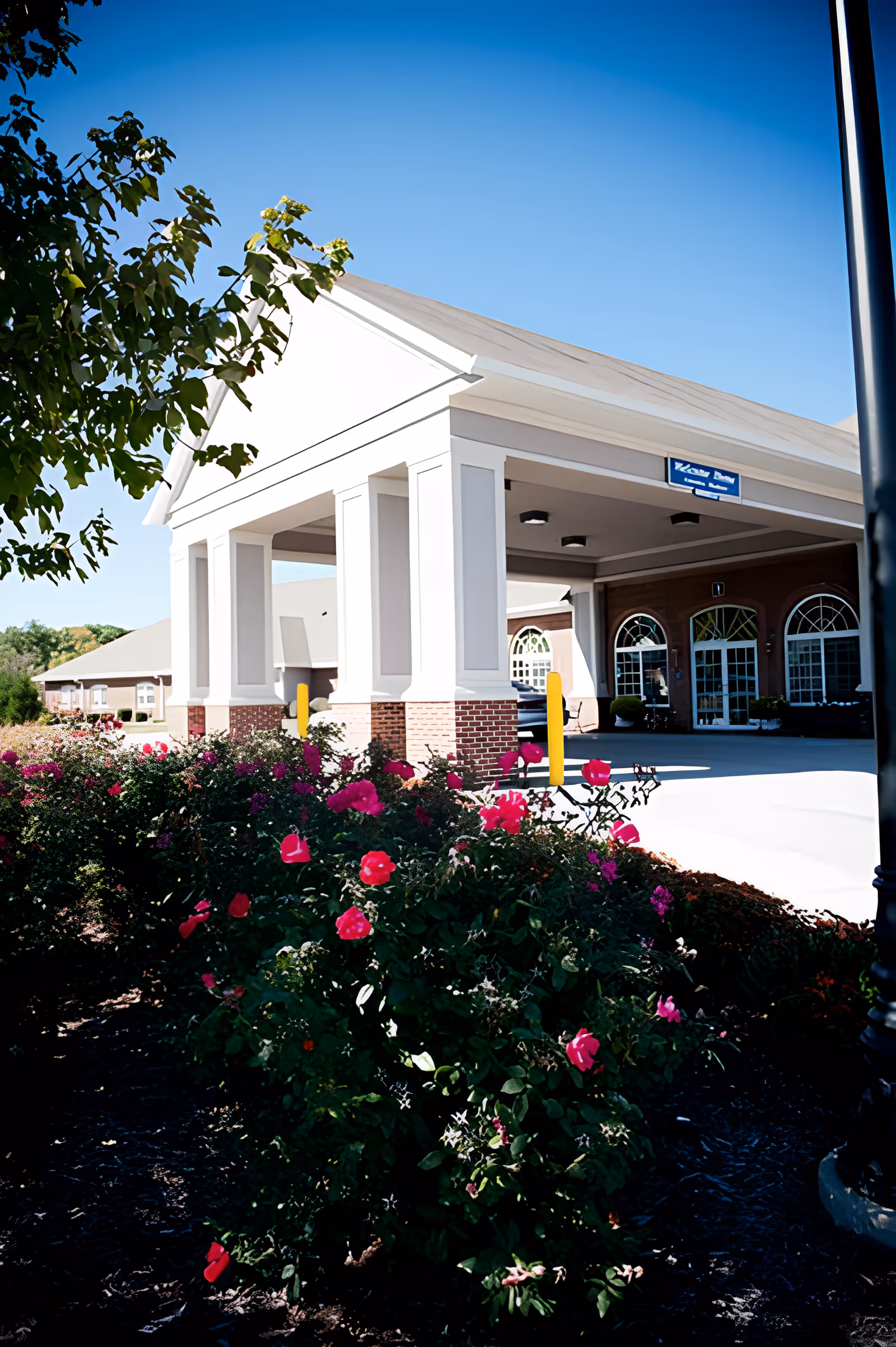 Exterior view of Coventry Meadows facility entrance with a covered drop-off area supported by white columns and brick bases. In the foreground, there are vibrant pink flowers and green bushes under a clear blue sky.