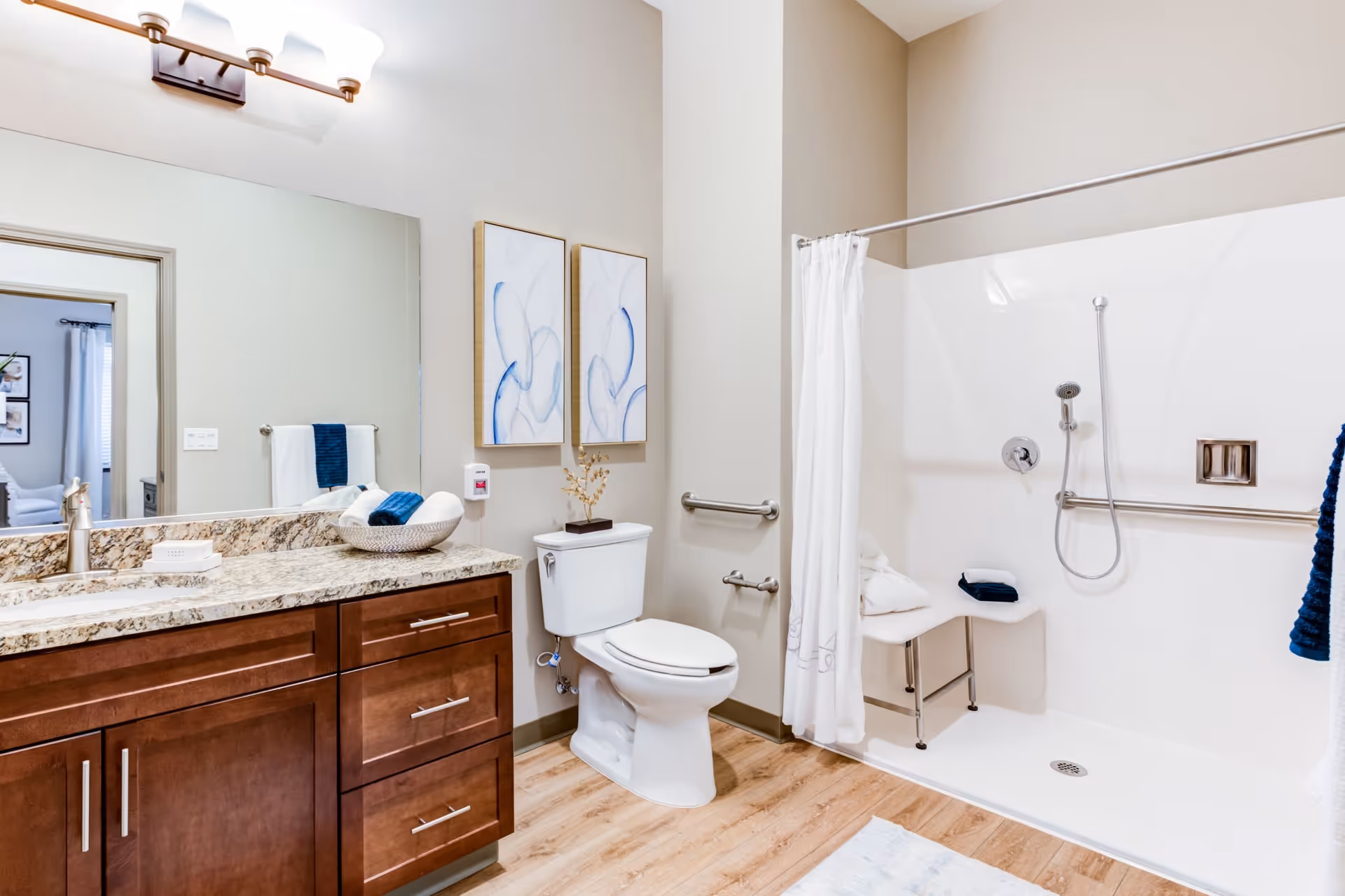 A clean and modern bathroom featuring a granite countertop with a sink, a large mirror, a toilet, and a walk-in shower with a white curtain and a built-in bench. The bathroom has light wood flooring and neutral-colored walls with two abstract blue and white paintings above the toilet.