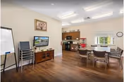 A well-lit common area with wooden flooring featuring a small kitchen area with cabinets, microwave, and popcorn machine. There is a table with chairs, a TV on a wooden stand, a stack of chairs, a whiteboard, and a framed picture on the wall.