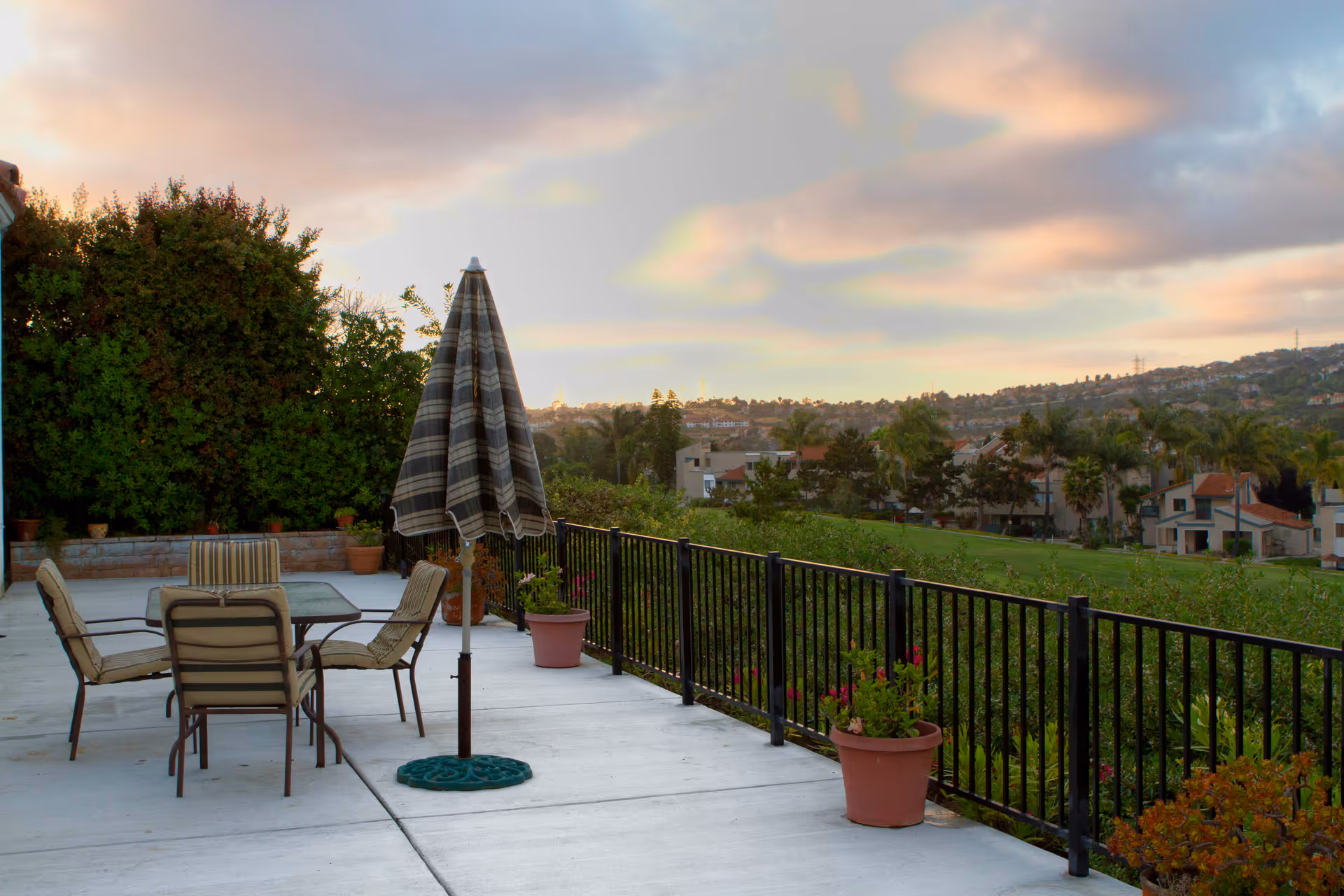 Outdoor patio area with a table and four cushioned chairs around it. A closed striped umbrella stands in the center of the table. Several potted plants line the edge of the patio, which is bordered by a black metal railing. In the background, there is a view of a green landscape with trees, houses, and hills under a partly cloudy sky during sunset.