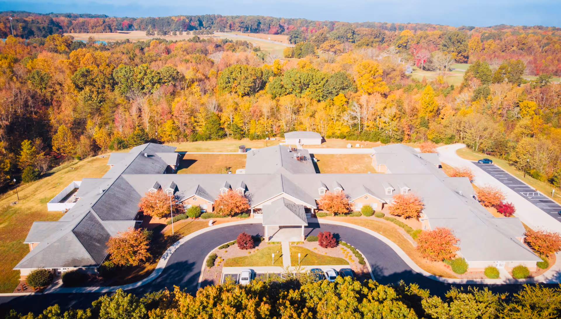 Aerial view of Caswell House, a large single-story building surrounded by trees with autumn foliage. The building has a U-shaped driveway with several parked cars and is set in a rural area with rolling hills and dense forest in the background.