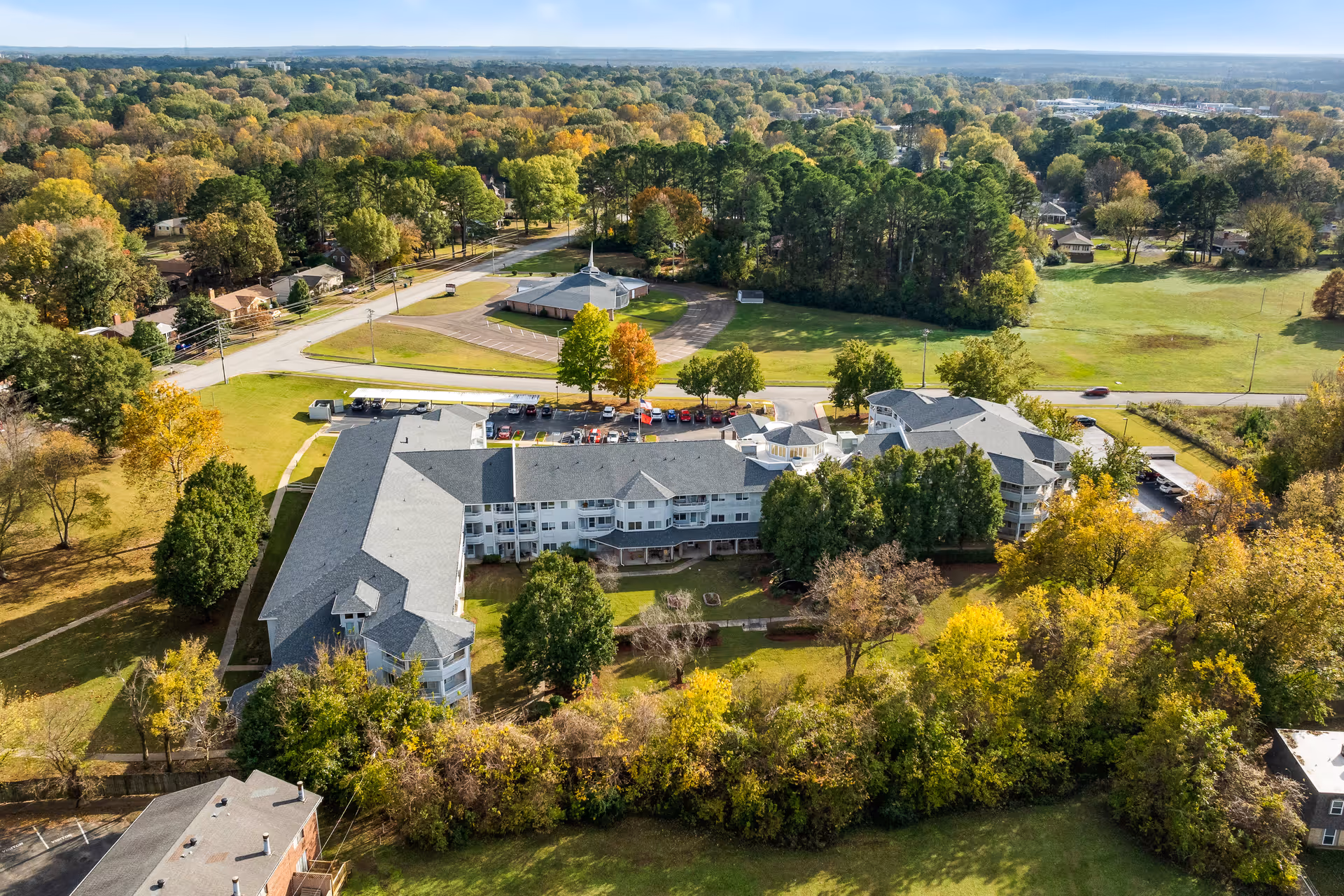 Aerial view of Jackson Meadow, a senior living community, showing a large multi-wing building surrounded by trees with autumn foliage. There is a parking lot with cars, open green spaces, and a road nearby under a clear blue sky.