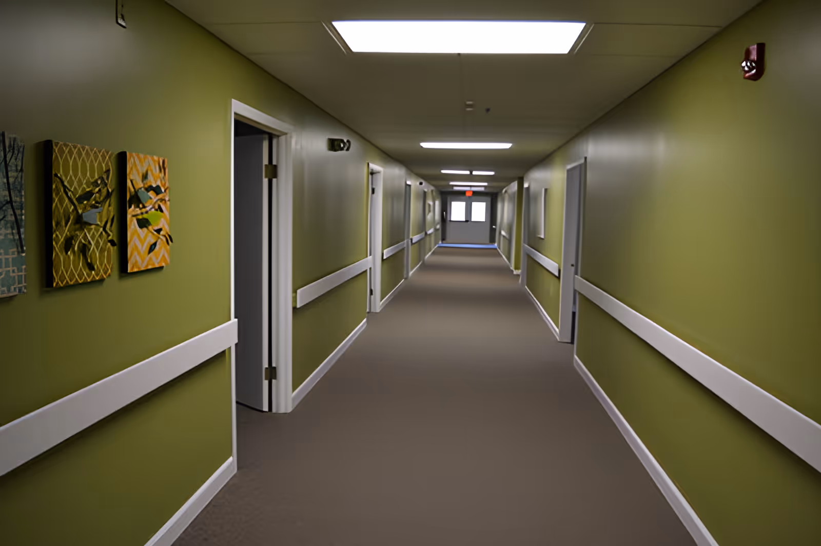 A long, empty hallway in a senior living facility with olive green walls, white handrails, and several doors on both sides. The ceiling has rectangular fluorescent lights, and there are three decorative wall hangings on the left side near the foreground.