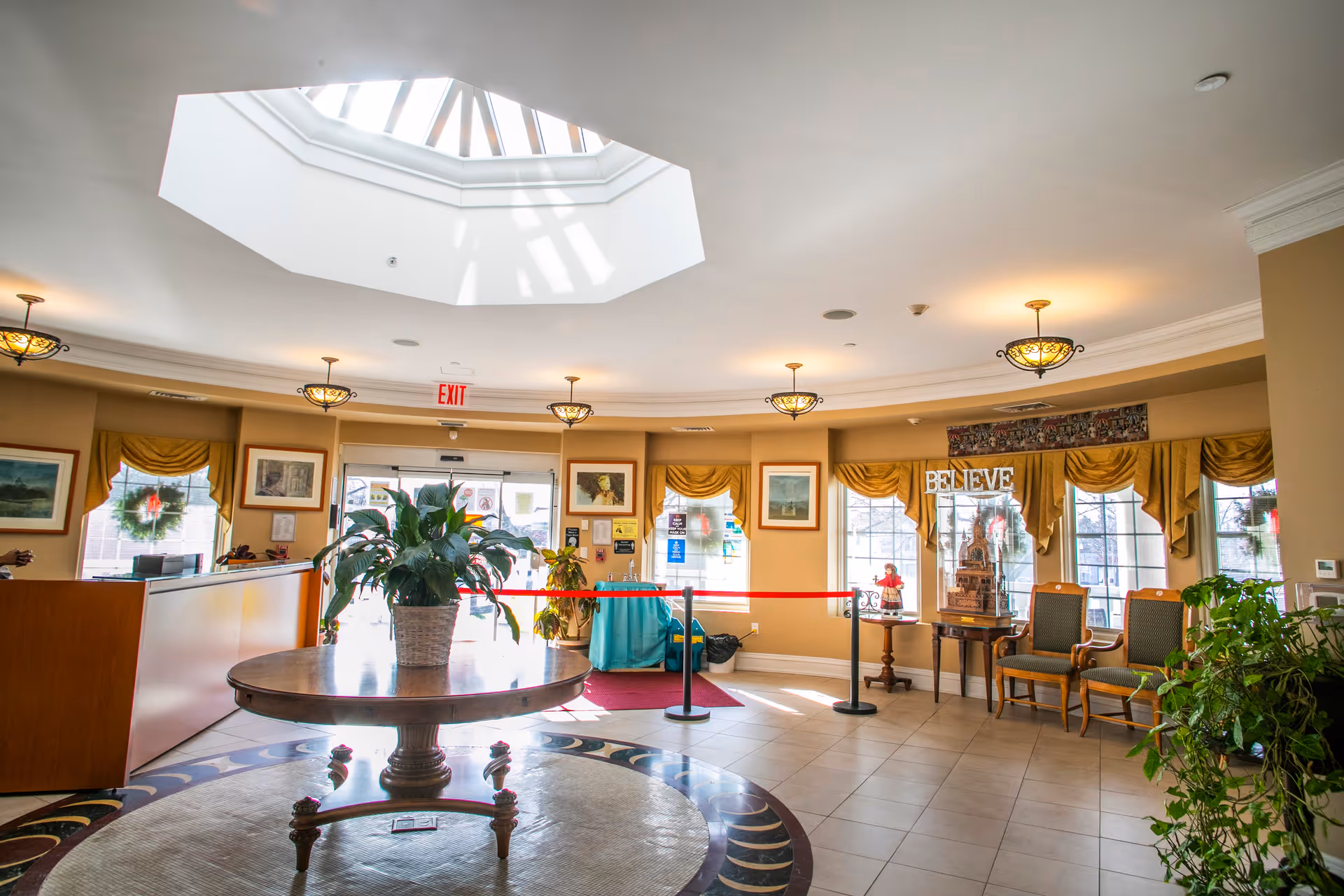 Bright and spacious lobby area with a large round wooden table holding a potted plant in the center. The room features a skylight ceiling, multiple hanging light fixtures, framed artwork on the walls, and large windows with yellow curtains. There are two wooden chairs with green cushions near a small table displaying a decorative piece with the word 'BELIEVE'. The entrance door is visible with an exit sign above it, and a red rope barrier is set up near the door.