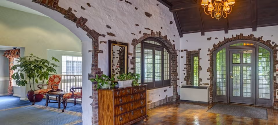 Interior view of a senior living facility lobby or common area with arched doorways and windows framed by exposed brick. The room features a wooden chest of drawers with potted plants on top, a chandelier hanging from a dark wooden ceiling, and a seating area with patterned chairs and a small table near a window.