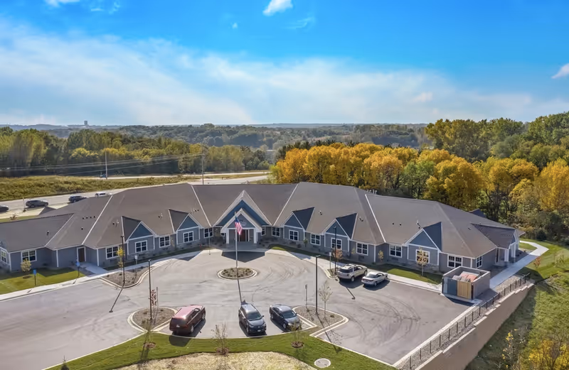 Aerial view of a single-story senior living facility with a circular driveway, parked cars, and surrounding trees.