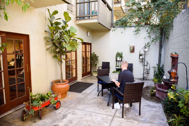 A man sitting at a table in a small outdoor courtyard area with potted plants, a spiral staircase, and a water fountain. The courtyard is surrounded by light-colored walls with glass doors leading inside the building.