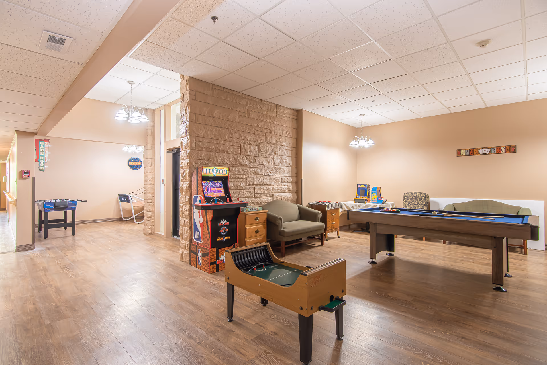 A spacious game room with wooden flooring and beige walls. The room features a pool table, foosball table, arcade basketball game, and other arcade machines. There are also several chairs and a small sofa arranged along the walls. The ceiling has white tiles with recessed lighting and hanging light fixtures. A sign on the wall reads 'GAME ROOM'.