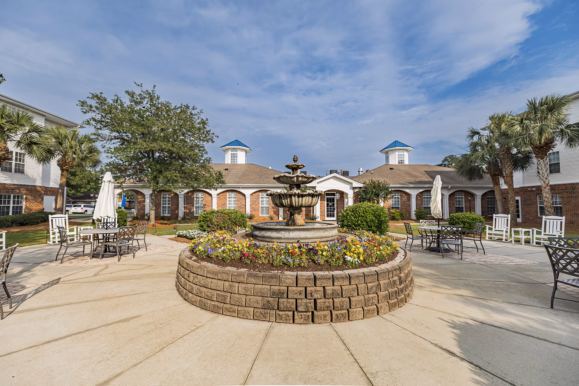 A courtyard with a tiered stone fountain surrounded by flowerbeds, patio tables, and a brick senior living building in the background.