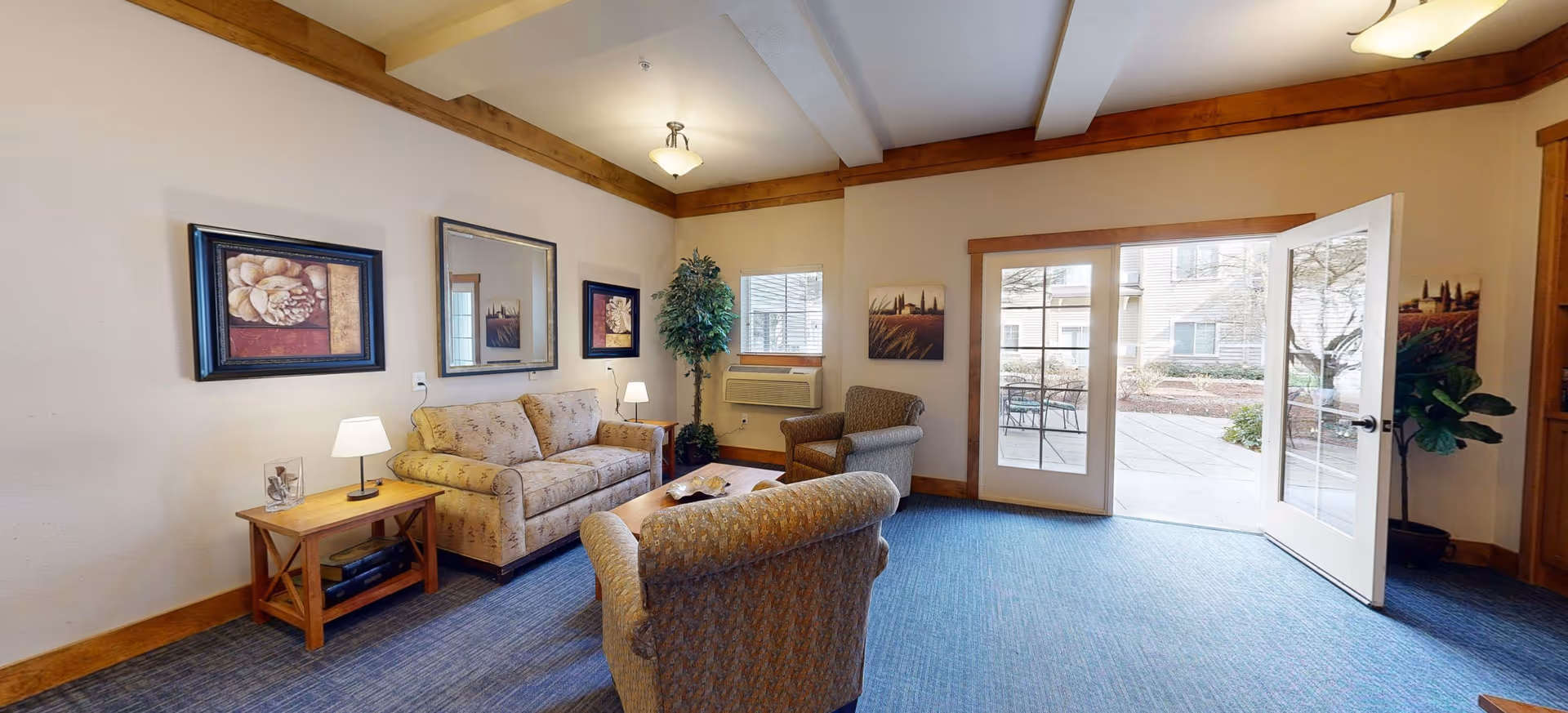 A cozy living room area with a beige patterned sofa, two armchairs, a wooden side table with a lamp, framed artwork on the walls, a large mirror, and a potted plant. The room has blue carpet flooring, cream-colored walls with wooden trim, and a glass door leading to an outdoor patio area.