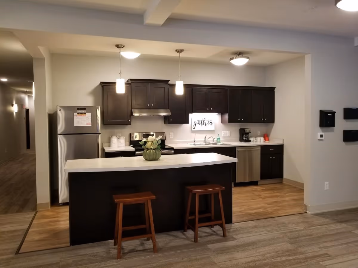 Modern kitchen area with dark wood cabinets, stainless steel refrigerator, stove, and dishwasher. A white countertop island with two wooden stools is in the foreground. Two pendant lights hang above the island, and a decorative sign that says 'gather' is mounted on the wall above the sink.