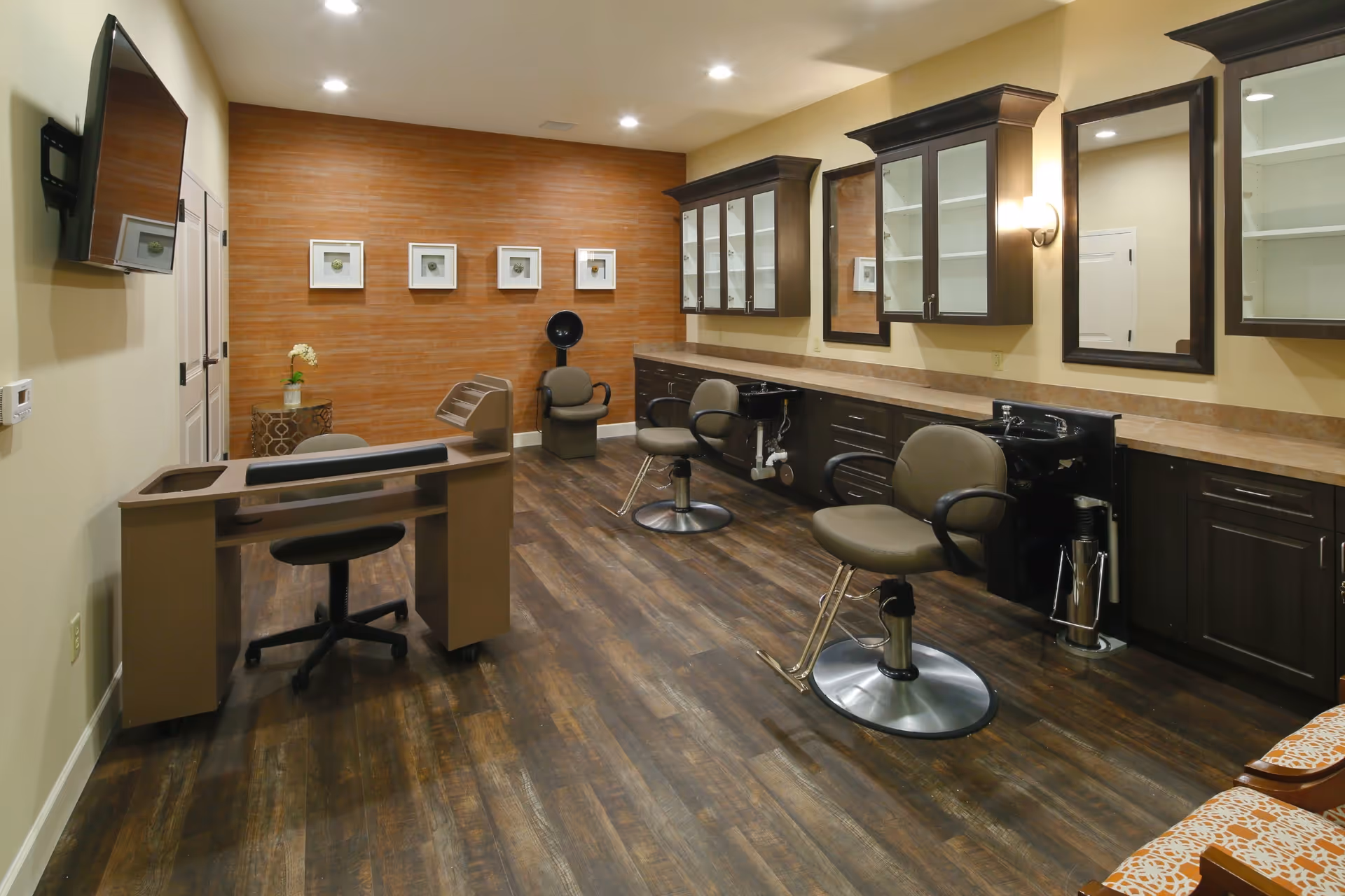Interior of a salon room with two salon chairs in front of mirrors and sinks, a hair dryer chair in the back, a manicure table with a rolling chair, wooden flooring, and a wall-mounted TV.