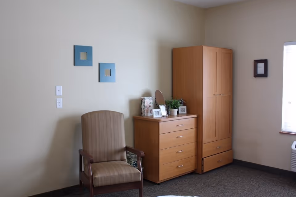 A simple room with an upholstered armchair, wooden dresser and wardrobe against a beige wall with small framed decorations and a window.