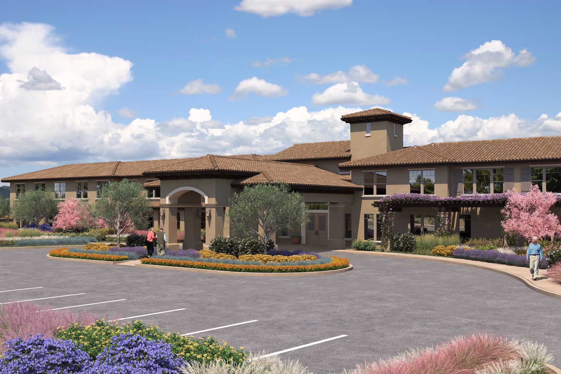 Exterior view of a senior living facility with a beige stucco building featuring a tiled roof, surrounded by landscaped gardens with colorful flowers and trees under a partly cloudy blue sky. There are a few people walking near the entrance and a parking area in the foreground.