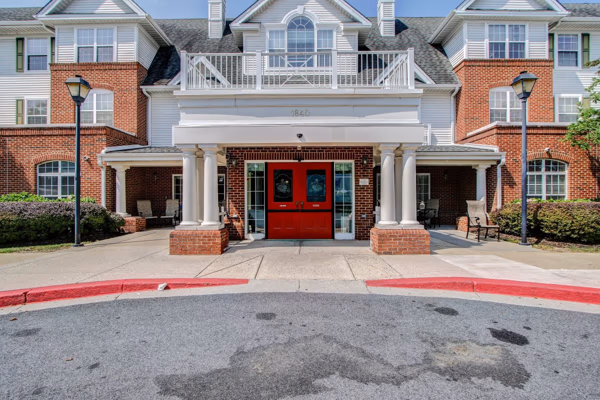 Front exterior view of a senior living facility with a brick and white facade, featuring a covered entrance with four white columns and double red doors. There are two lamp posts on either side of the entrance and some outdoor seating areas with chairs. The building has multiple windows and a peaked roof with dormers.