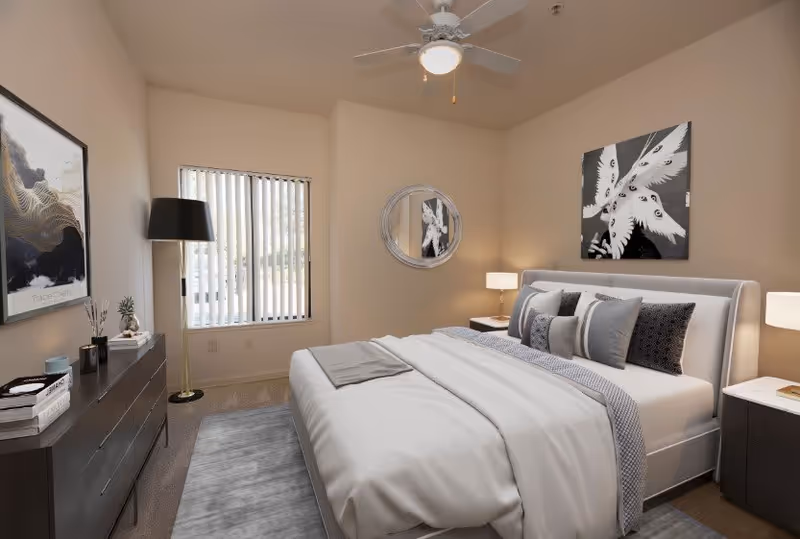 A neatly arranged bedroom with a large bed featuring multiple pillows and a white comforter. The room has beige walls, a ceiling fan with a light, a window with vertical blinds, a round mirror on the wall, two bedside tables with lamps, a dark dresser with decorative items, and two pieces of modern artwork on the walls.