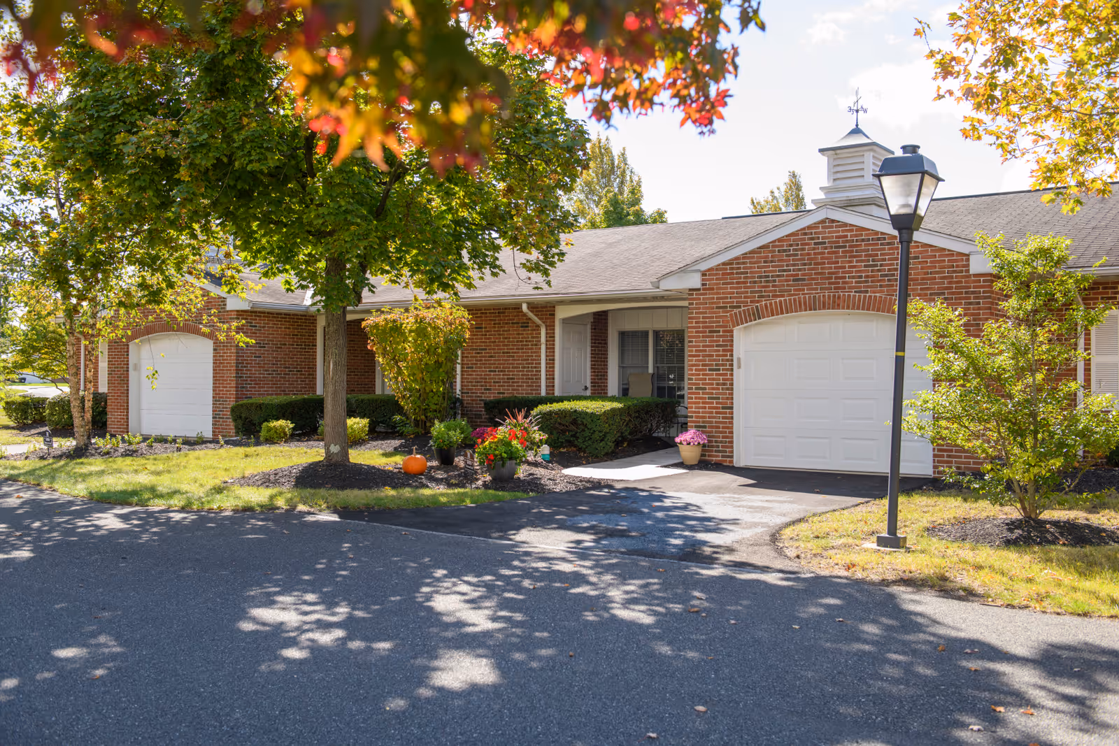 Exterior view of a single-story brick building with white garage doors and a small front porch. The area is landscaped with green bushes, a tree with some autumn-colored leaves, potted flowers, and a pumpkin near the entrance. A black street lamp stands near the driveway under a clear sky.