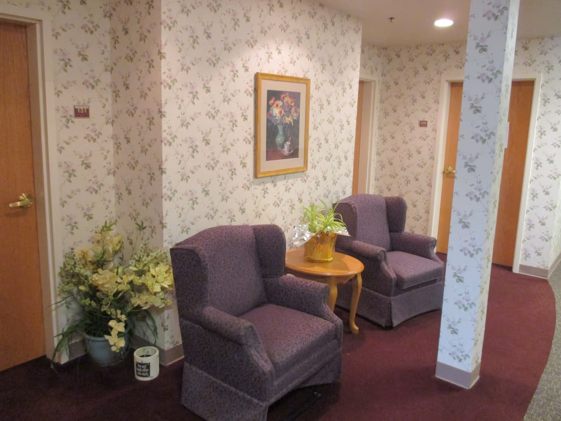 A hallway in a senior living facility with floral wallpaper and maroon carpet. Two purple upholstered armchairs are placed around a small wooden table with a potted plant on it. There is a framed picture of flowers on the wall above the chairs. Several wooden doors with room numbers are visible along the hallway, and a large potted plant is placed in the corner.