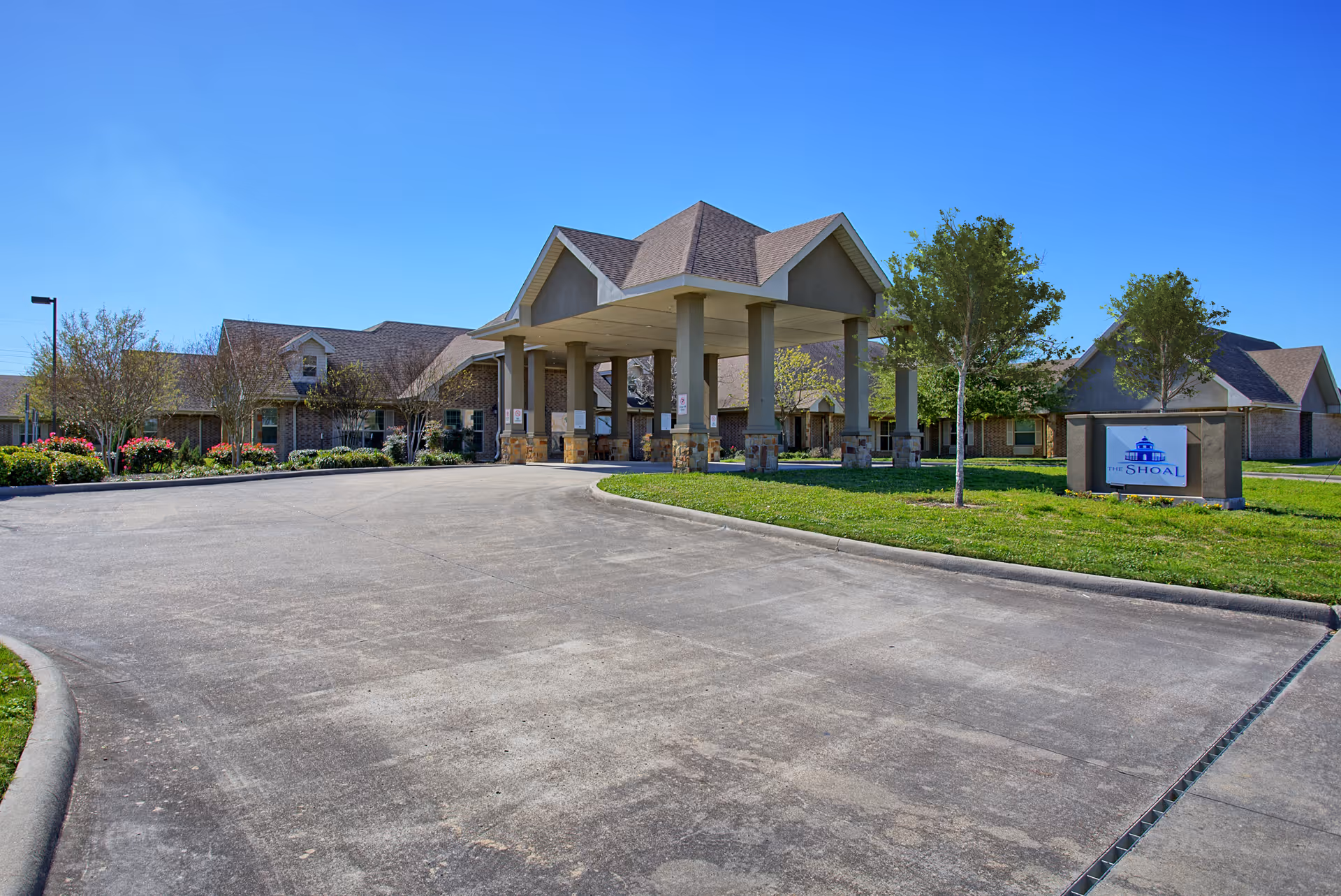 Exterior view of The Shoal senior living facility showing a large covered entrance with multiple pillars, surrounded by landscaped greenery and trees under a clear blue sky.