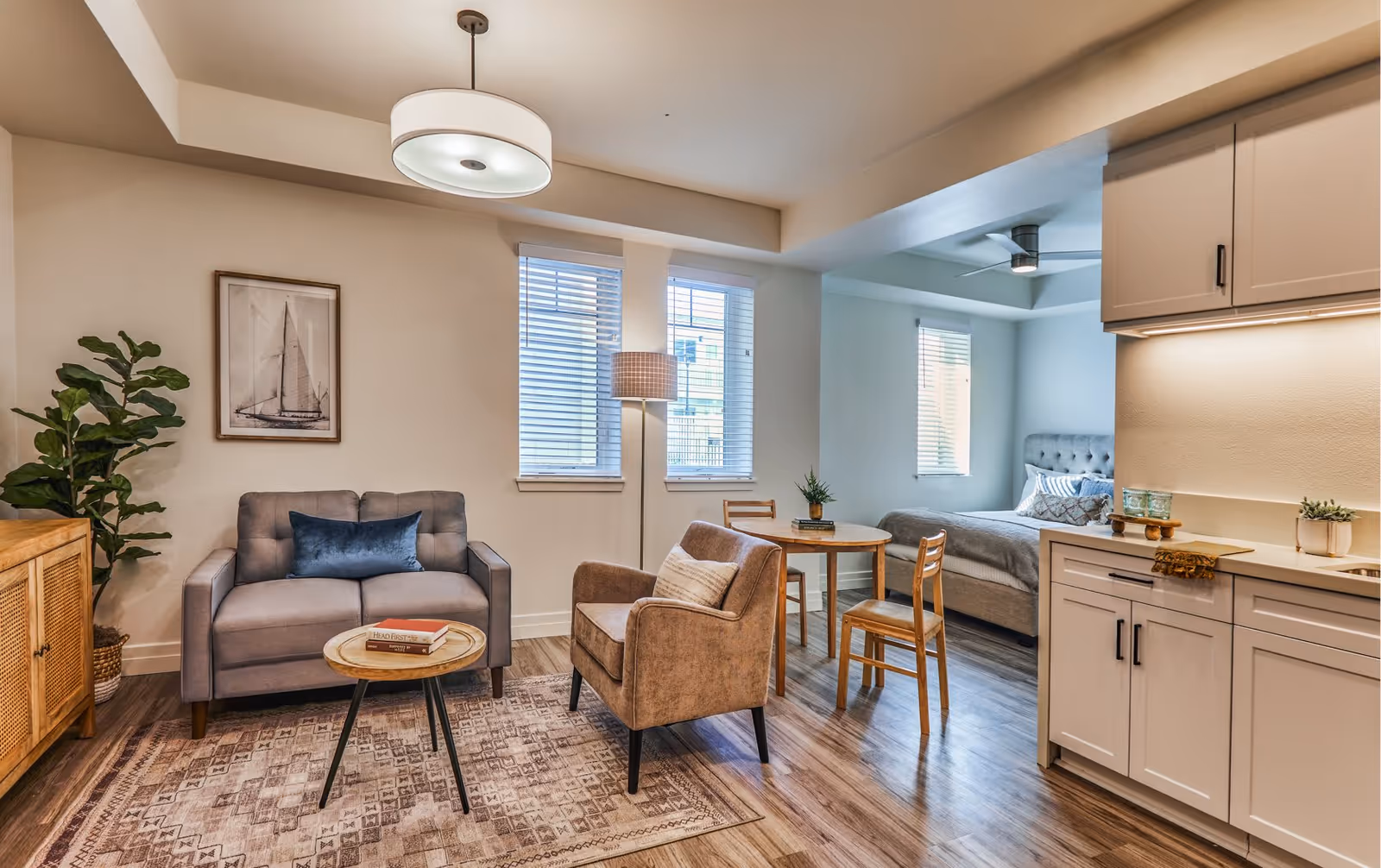 Furnished studio apartment interior with a sofa and armchair around a coffee table, a small dining table, kitchenette cabinets, and a bed in an adjacent alcove.