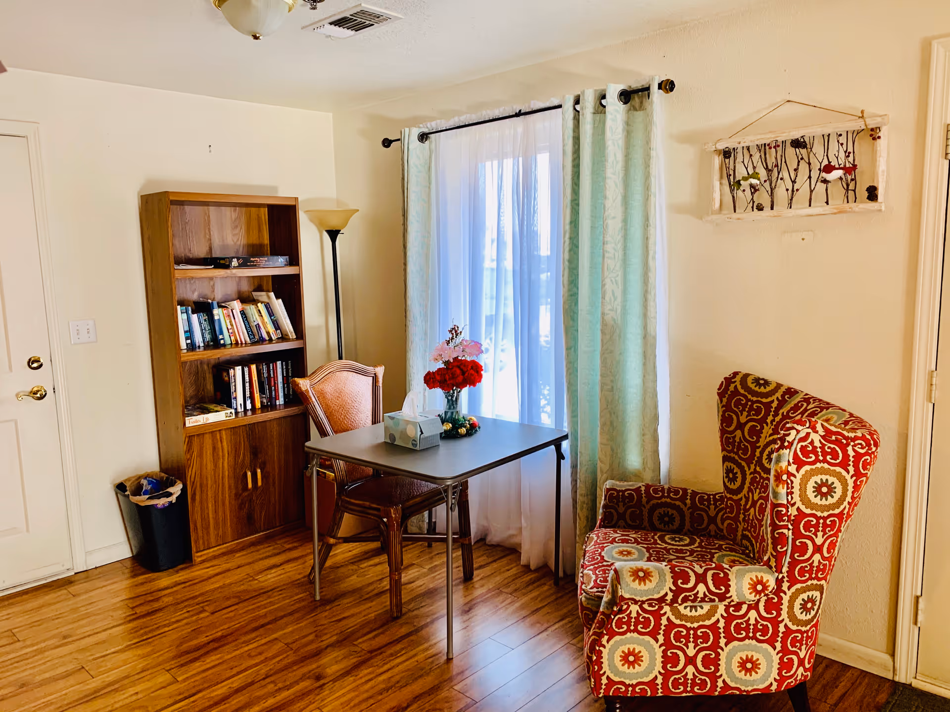 A cozy interior room with wooden flooring featuring a small table with a tissue box and a vase of flowers, a wooden bookshelf filled with books, a floor lamp, a patterned armchair, and a window with sheer white curtains and green drapes.