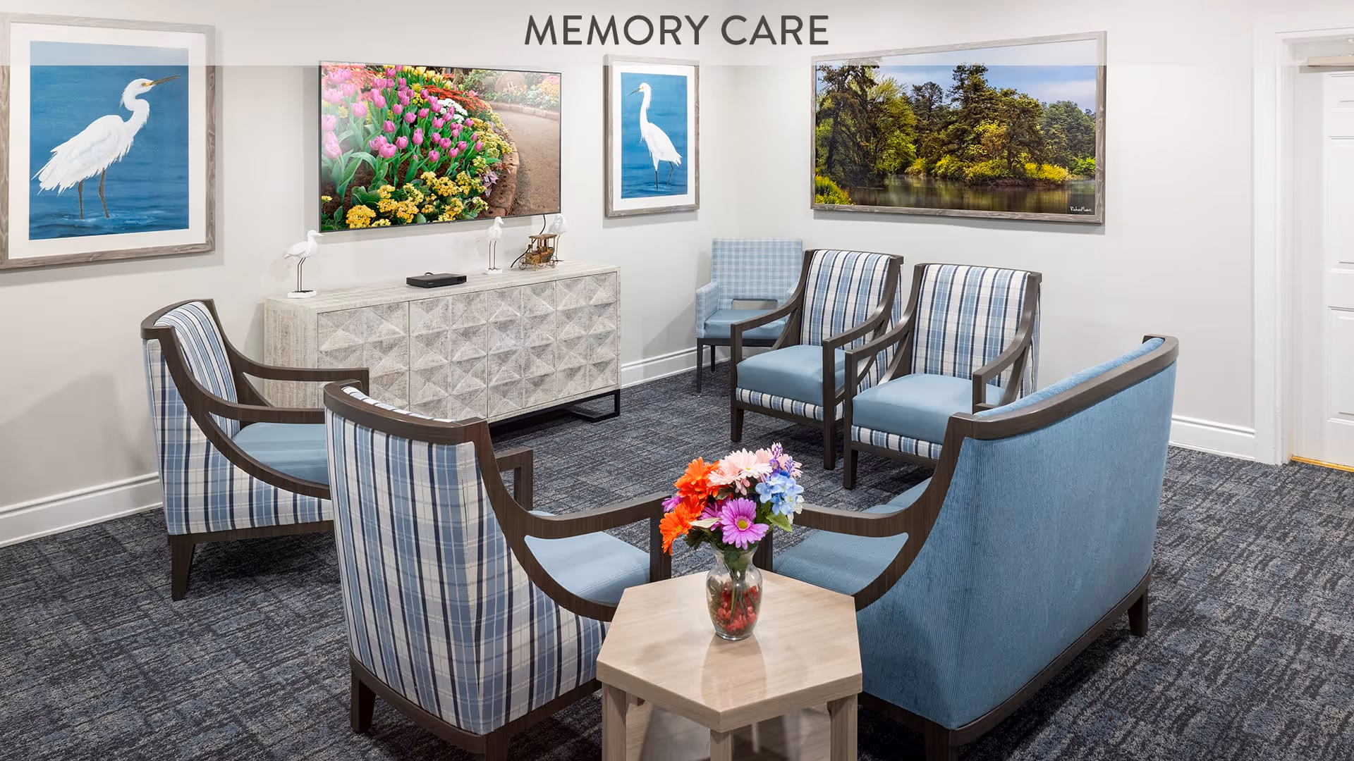 A cozy memory care seating area with six blue and plaid upholstered chairs arranged around a small wooden table with a vase of colorful flowers. The room has a patterned carpet and light-colored walls adorned with framed pictures of birds, flowers, and a scenic landscape. A white cabinet with decorative items sits against the wall under the 'MEMORY CARE' sign.