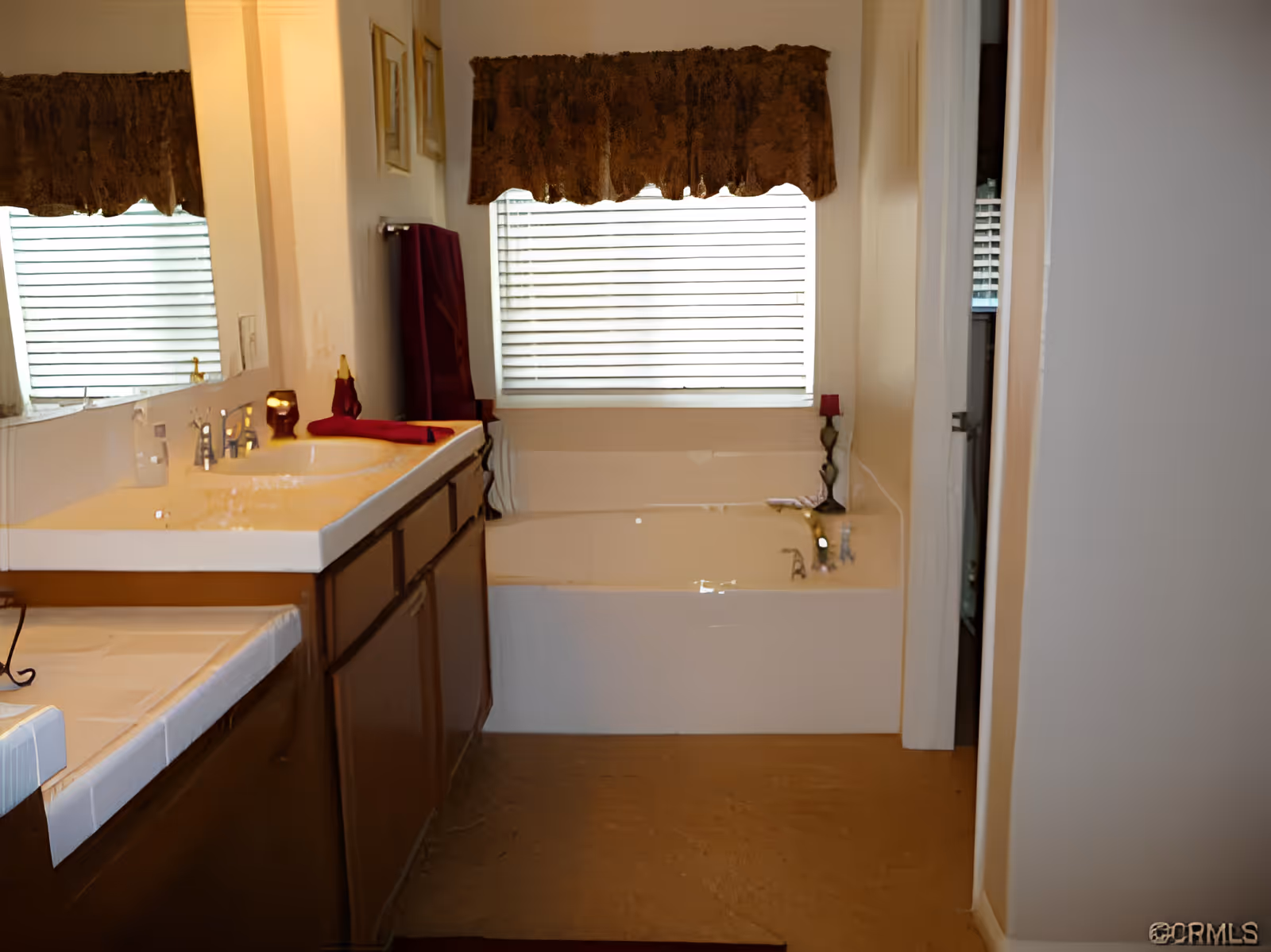 Bathroom with a large mirror above a double sink vanity with brown cabinets. There is a window with blinds and a brown valance above a white bathtub. A red towel and decorative items are placed on the vanity and bathtub edges.
