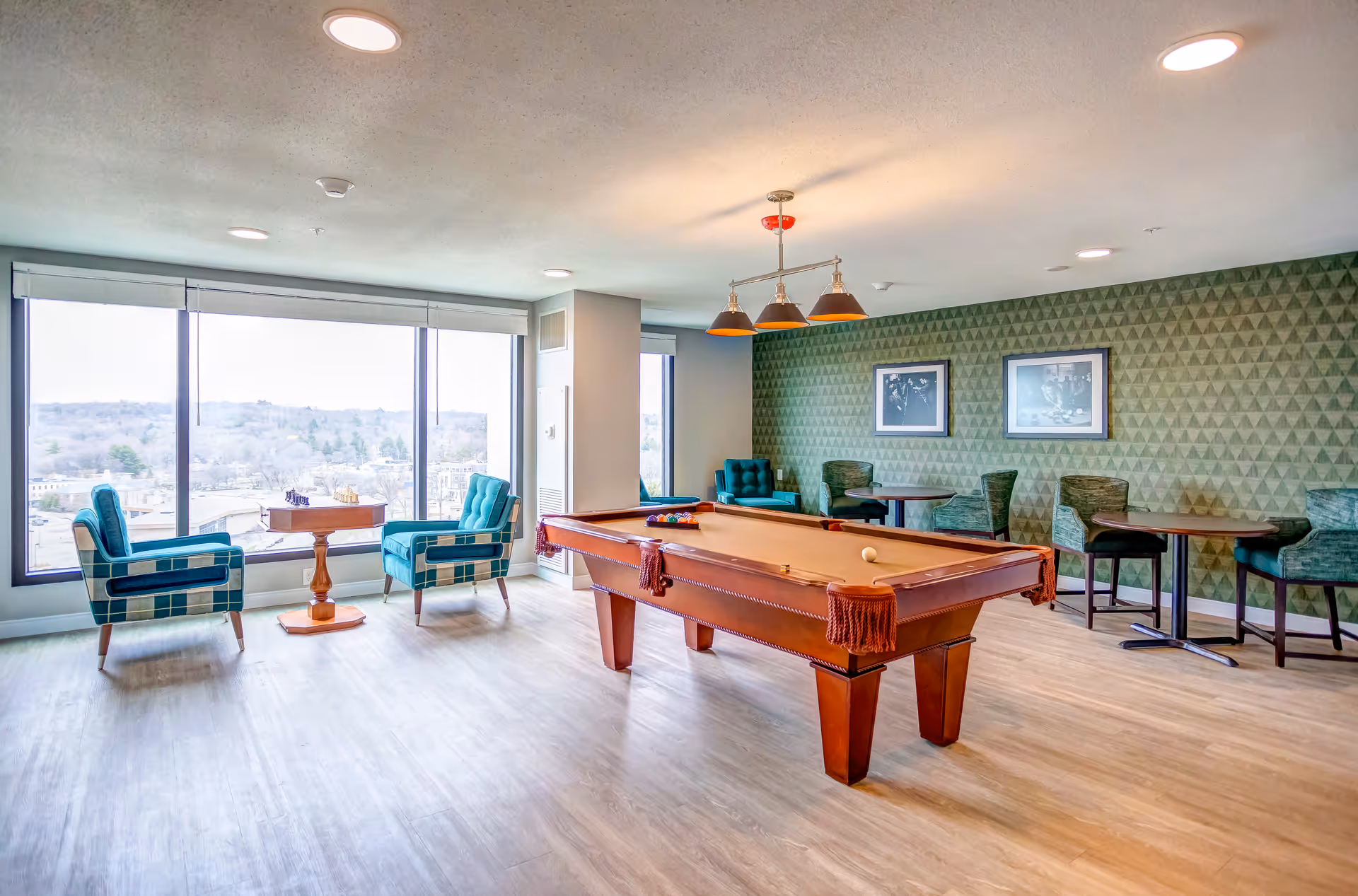 A bright recreational room with a pool table in the center, two teal armchairs with a small wooden table between them near large windows overlooking a scenic outdoor view, and several small round tables with chairs along a green patterned wall decorated with framed pictures.
