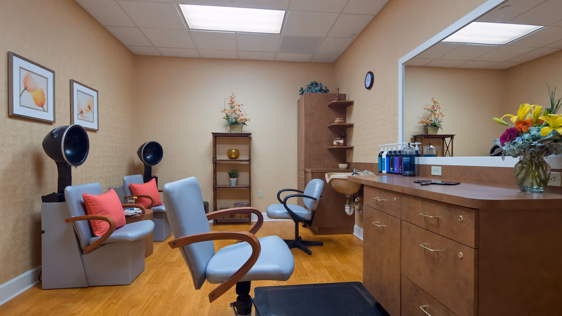 Interior of a hair salon area in a senior living facility with two hair drying chairs with black hooded dryers, two salon chairs with wooden armrests, a large mirror above a wooden counter with hair care products, a small sink, and decorative flowers and plants on shelves and the counter.