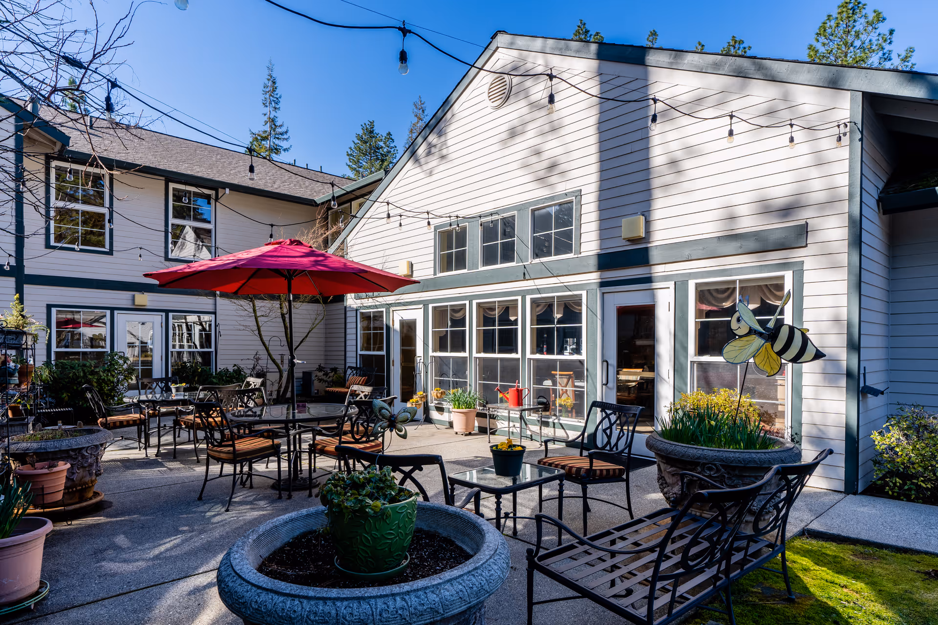 Outdoor patio area at Cascades of Grass Valley featuring metal tables and chairs with cushions, a large red umbrella, potted plants, string lights overhead, and a decorative bee sculpture near the building entrance.